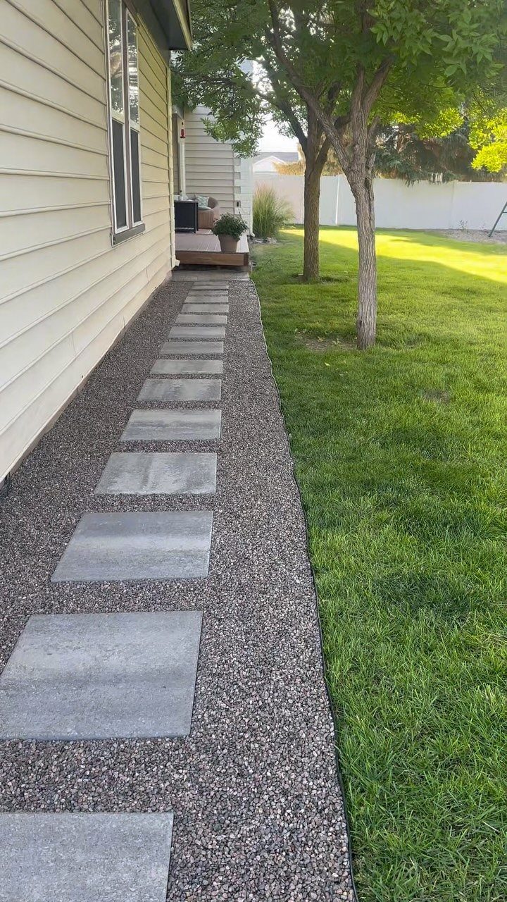 Rectangular grey stepping stones set into a narrow gravel path alongside a house and lawn.