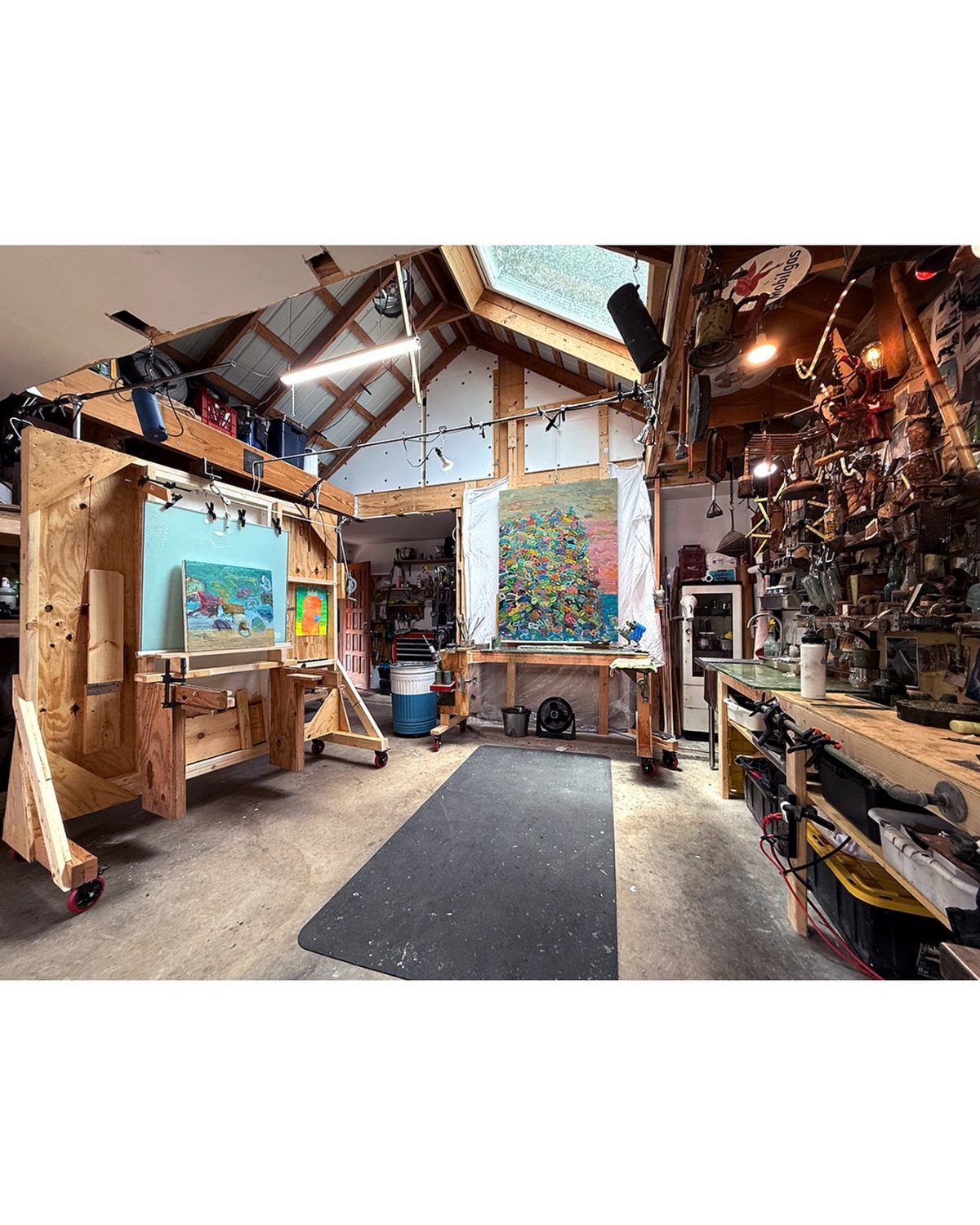An art studio inside a garage featuring easels, colorful paintings, and various art supplies on wooden workbenches.