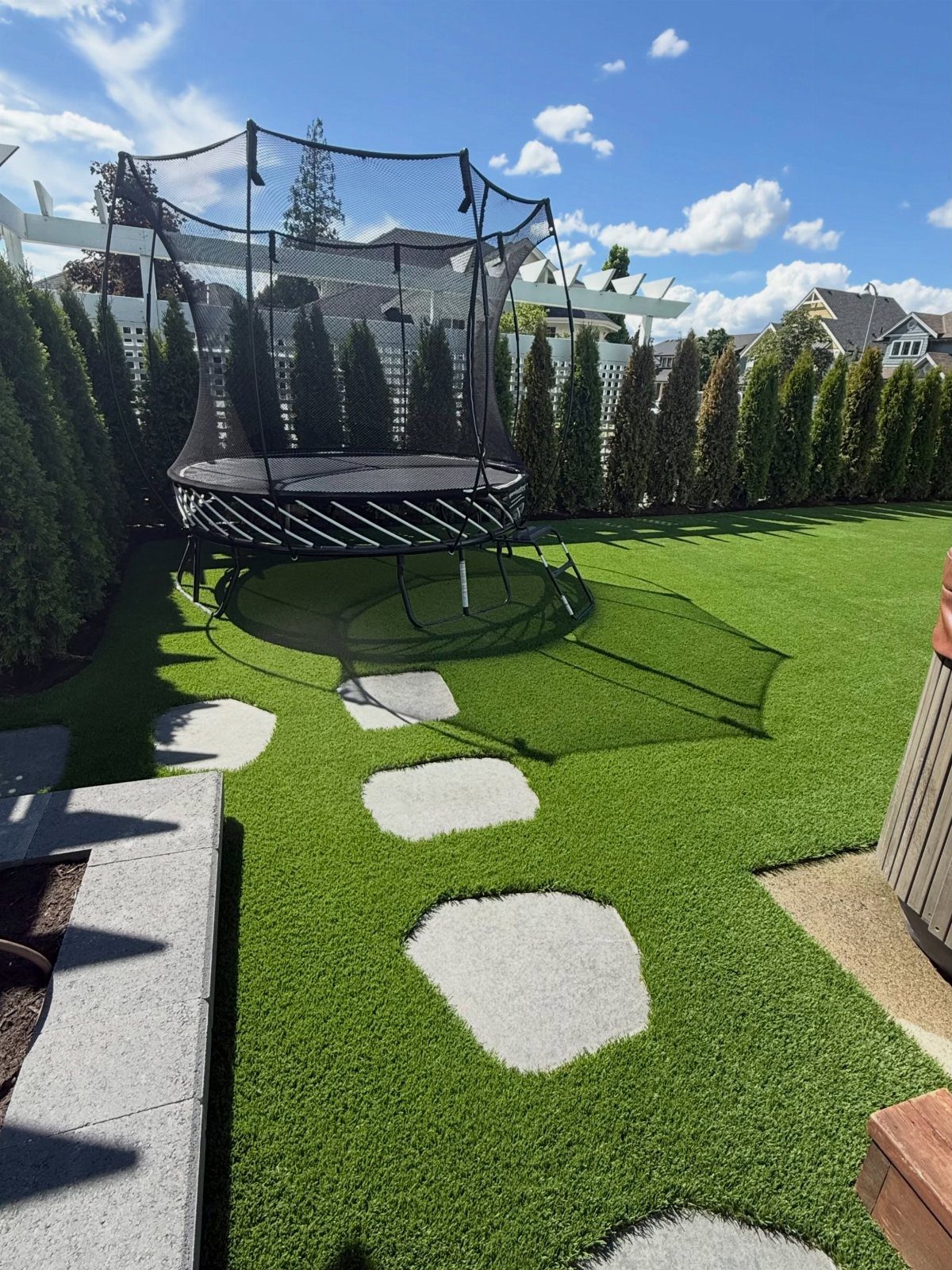 A black trampoline on a bright green artificial turf lawn with stepping stones and a hedge.
