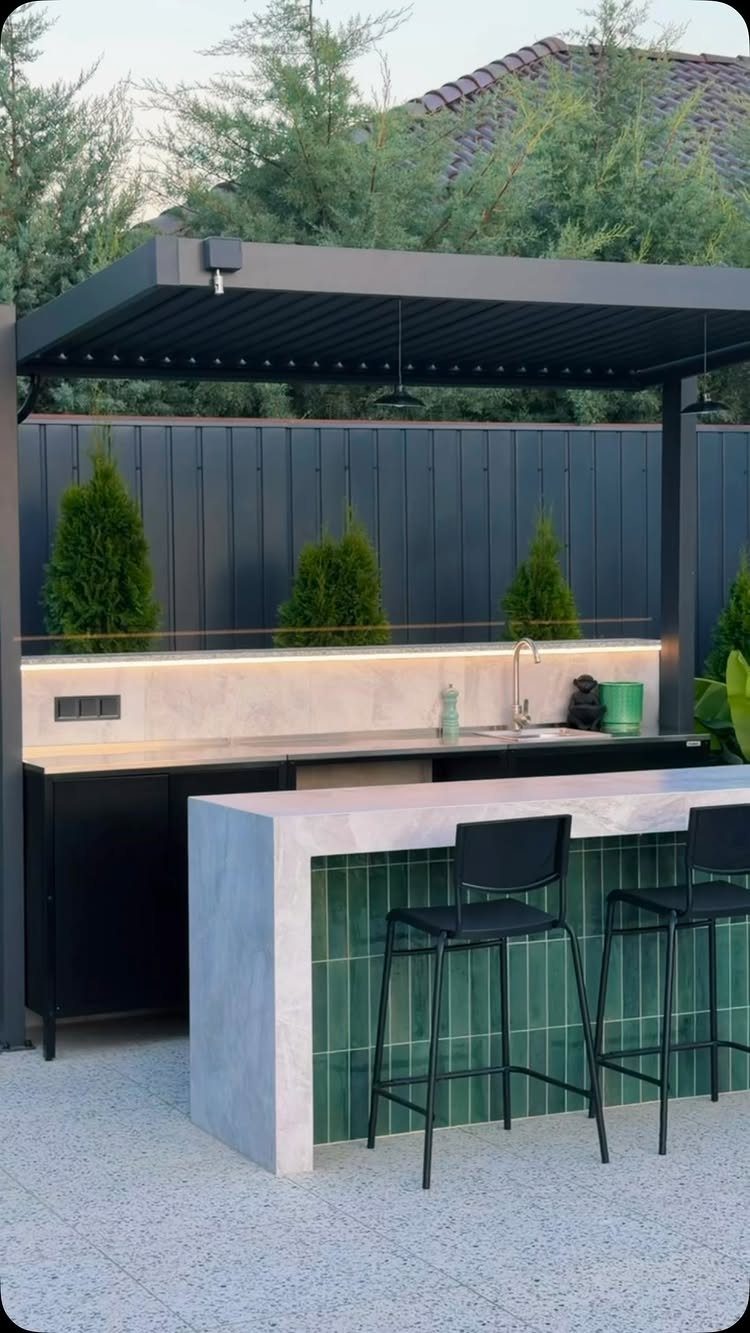 Modern outdoor beverage bar with stone countertop, green tiles, and black bar stools under a pergola.
