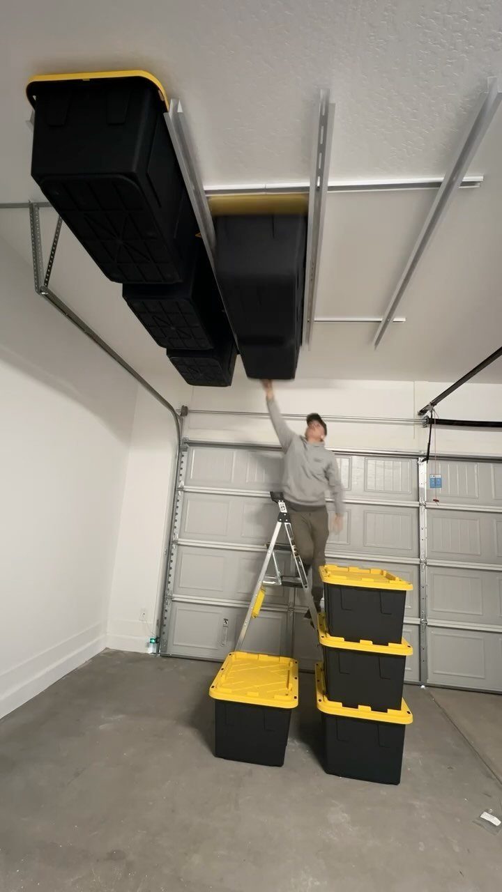 A man on a ladder reaches for black storage bins hanging from ceiling tracks in a garage.