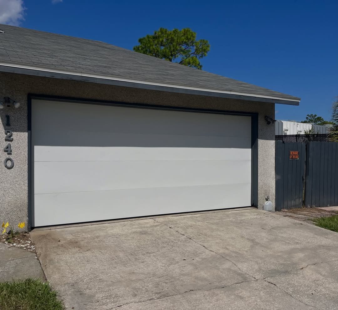 A white garage door with clean flush panels sits under a grey roof on a sunny day.