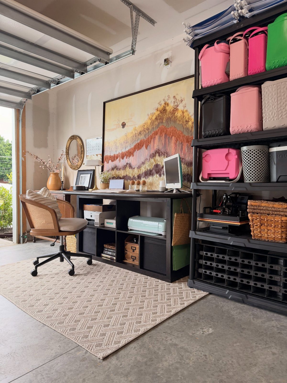 Organized craft room workspace with a black desk, computer, colorful storage bins, and a large landscape painting.
