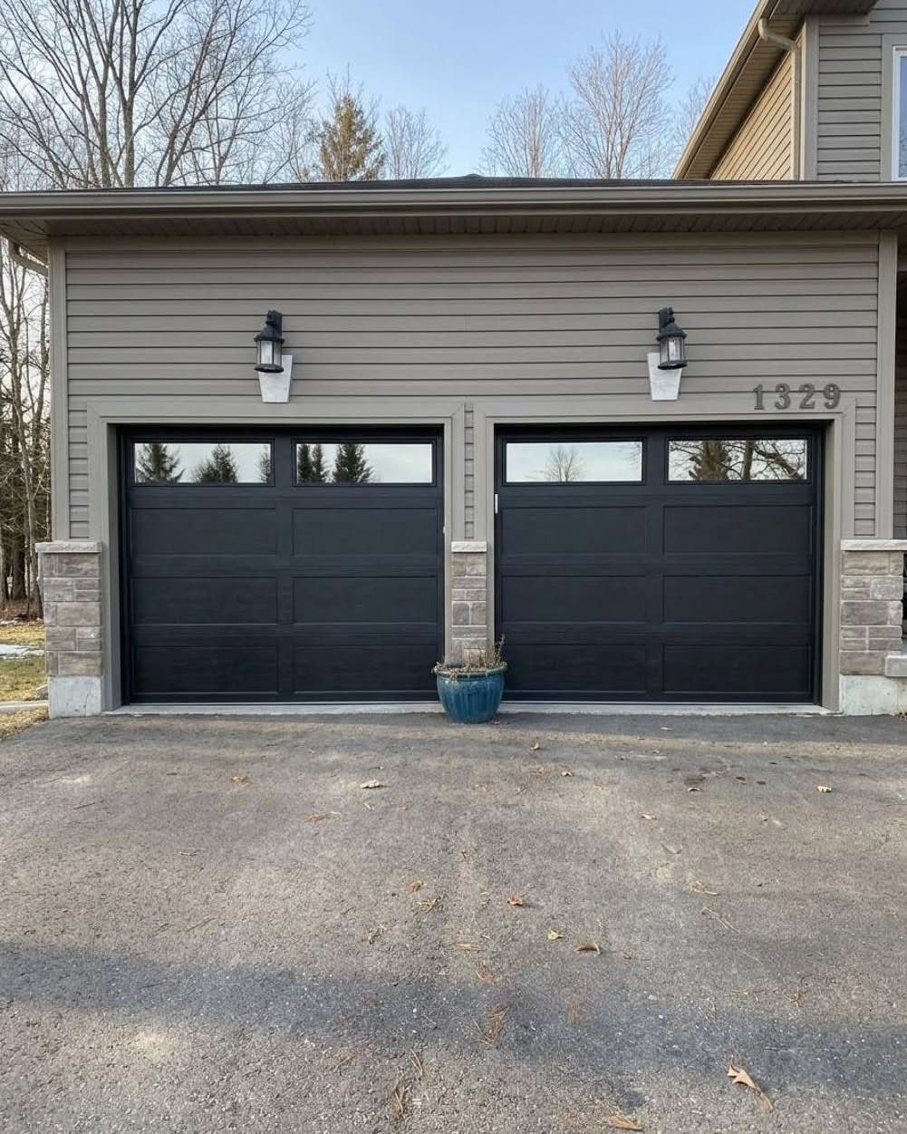 Two black modern garage doors with windows on a grey house with outdoor wall lanterns.