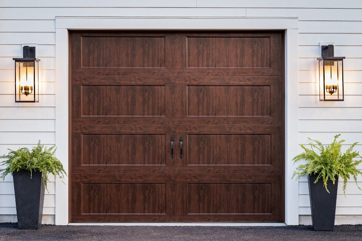 Dark wood garage door with black handles and lantern lights on white siding.