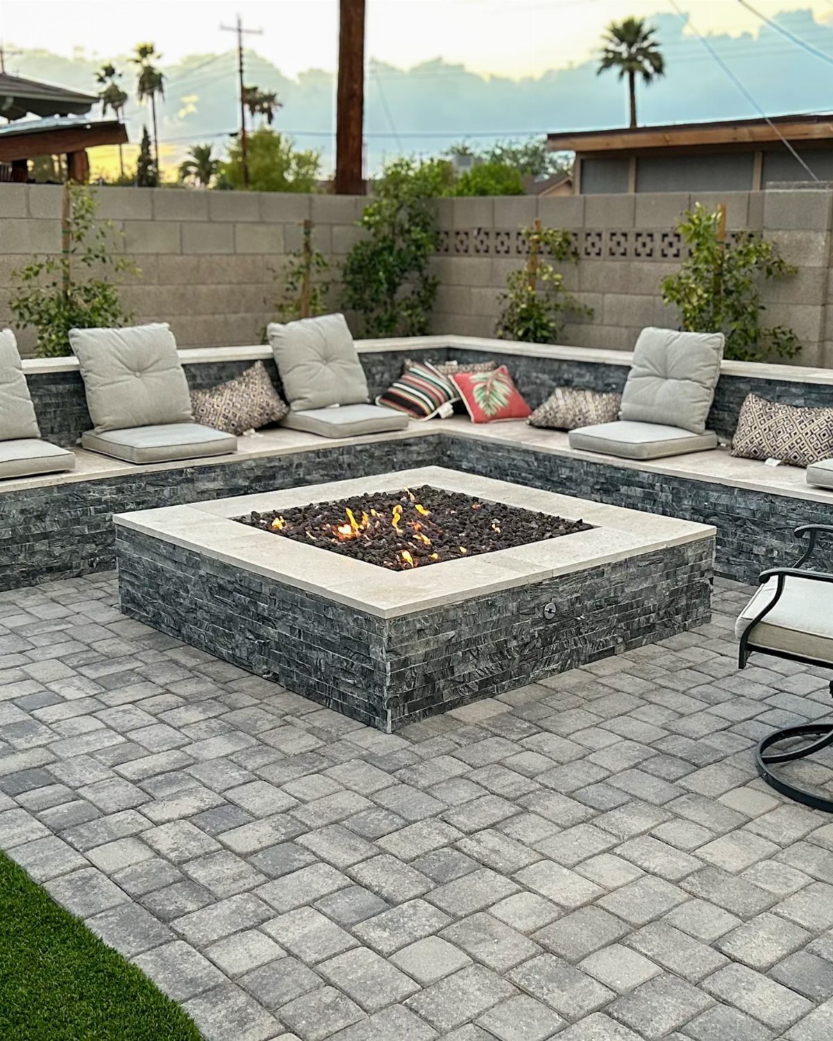 A square stone fire pit with glowing embers on a grey paved patio.