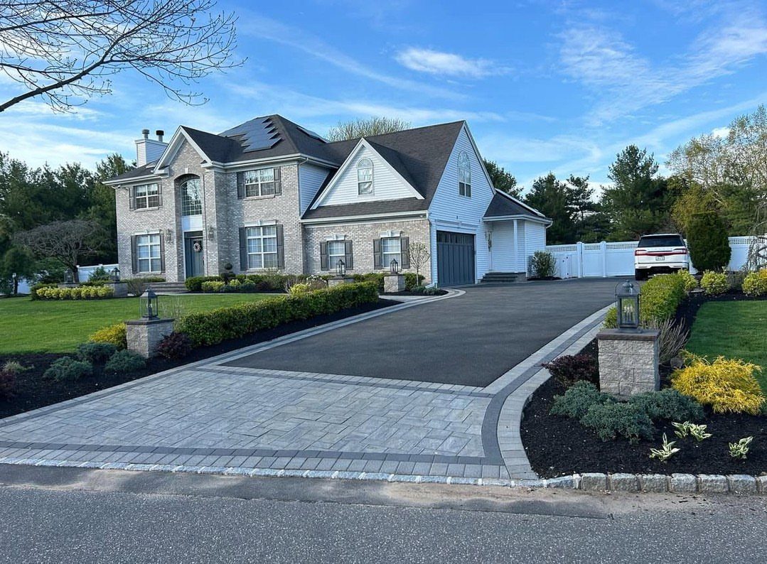 A large grey stone house featuring a paved stone driveway entrance and manicured landscaping.