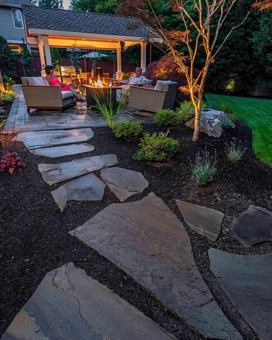 11 Flagstone Patio Design Ideas with Photos for Your Yard 3 Irregular flagstone stepping stones embedded in dark mulch leading toward a lit outdoor patio area.