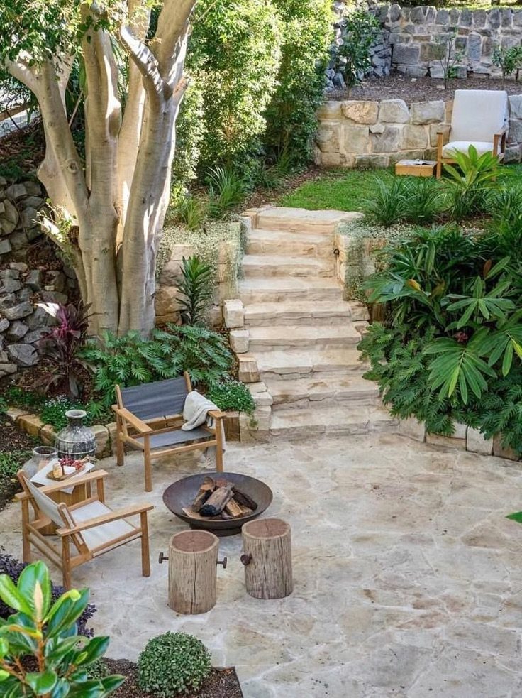 A flagstone patio with stone steps, a fire pit, and wooden chairs in a lush garden.