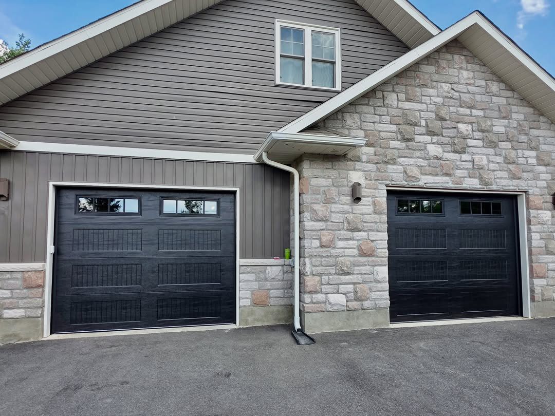 Two black garage doors with small windows on a house with stone and grey siding.