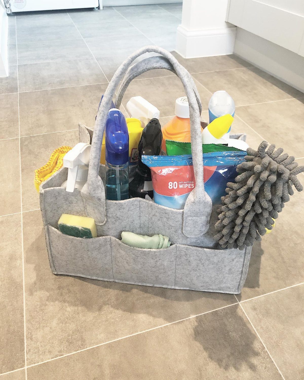 A grey felt caddy filled with various cleaning supplies sitting on a tiled floor.