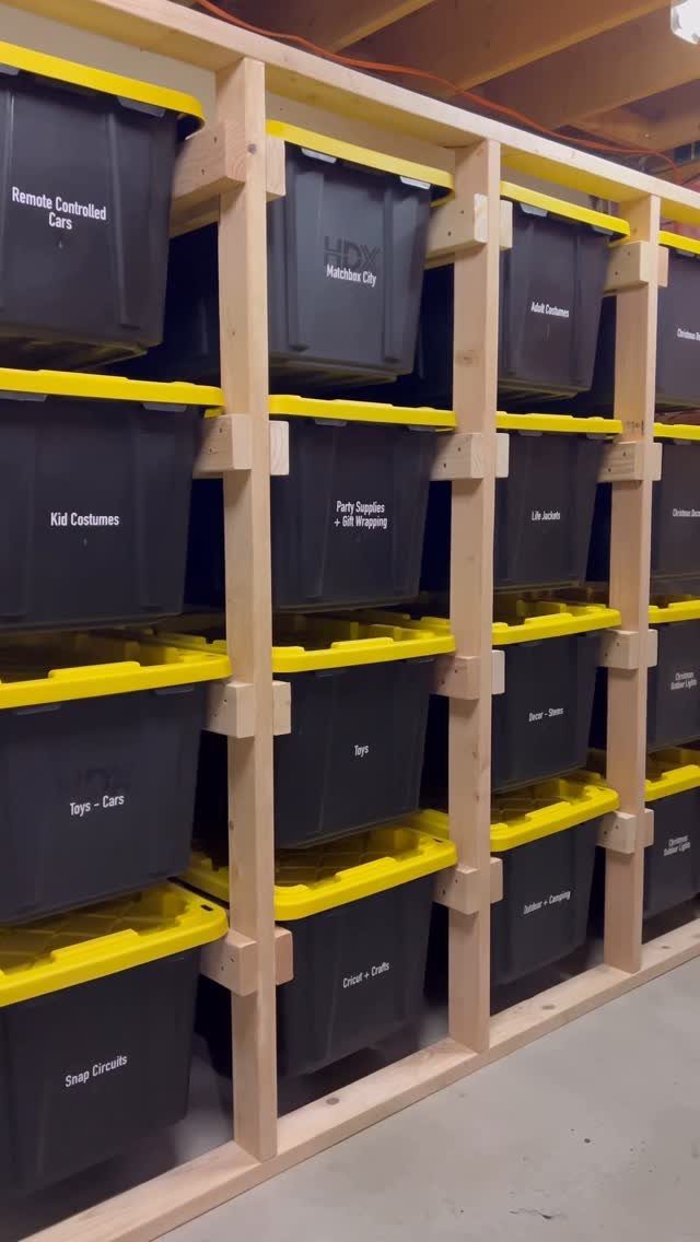 Black storage bins with yellow lids organized on a wooden shelving unit in a garage.