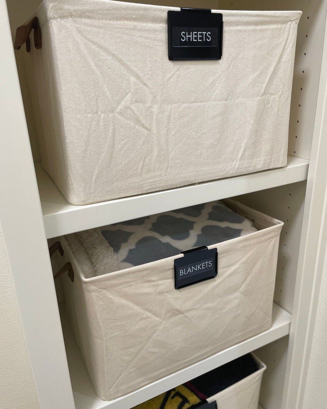 DIY Closet Organization: 16 Ideas for Every Layout 9 White fabric storage bins with black labels for sheets and blankets on white closet shelves.