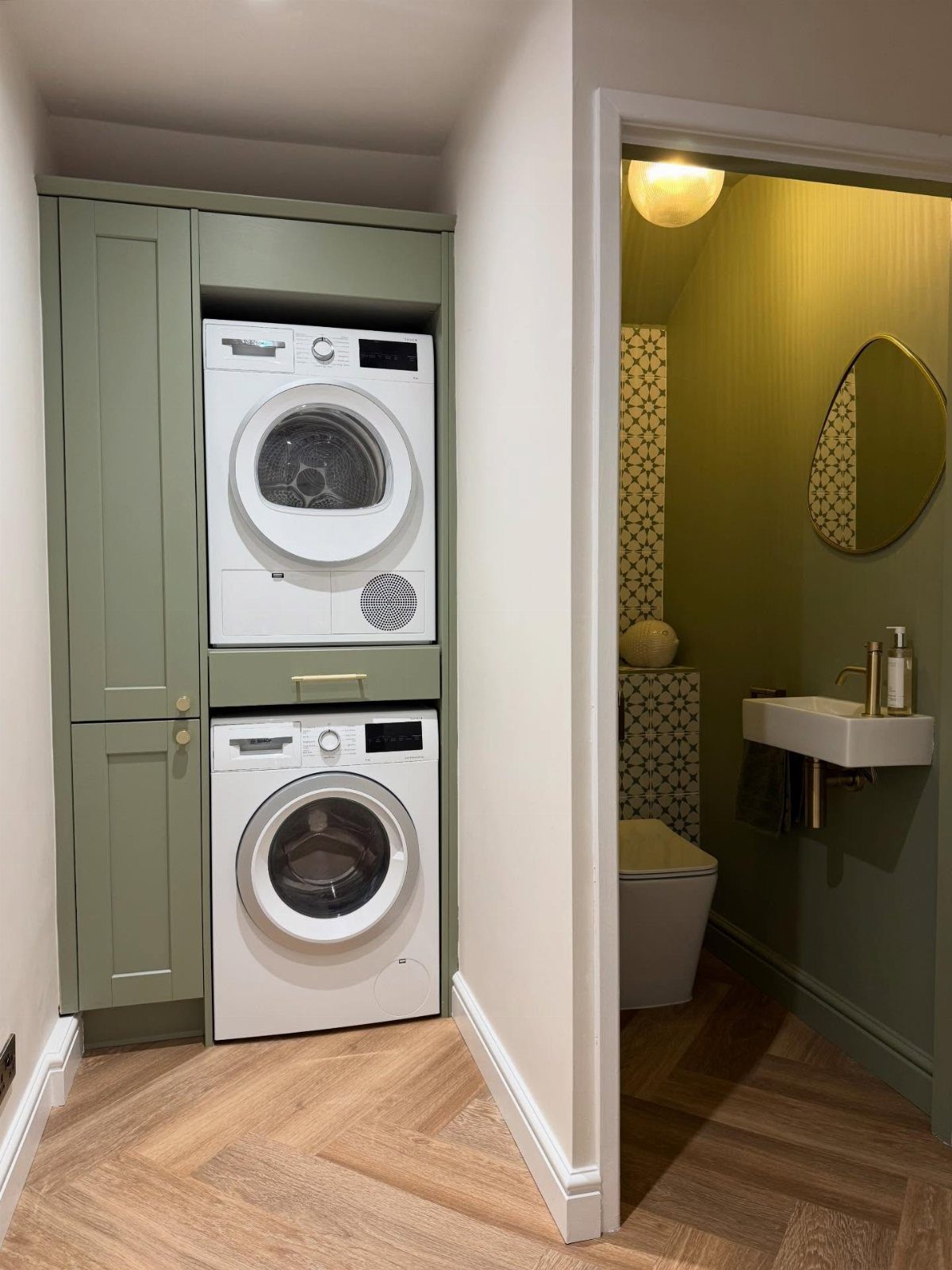 Stacked white laundry machines built into sage green cabinetry next to a small bathroom doorway.