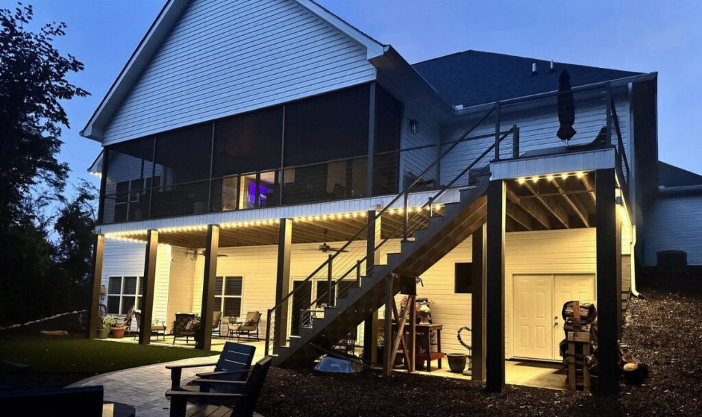 A lit patio area with furniture situated directly underneath a large elevated wooden deck at dusk.