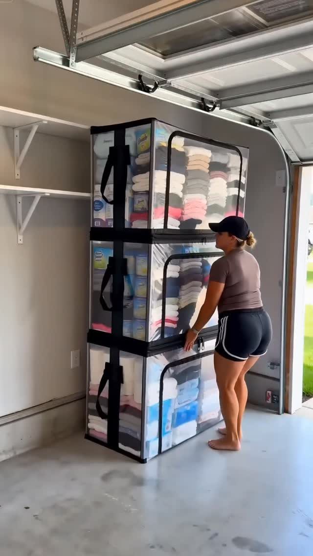 A woman stands beside three large, clear, stackable fabric storage bins filled with folded clothes in a garage.