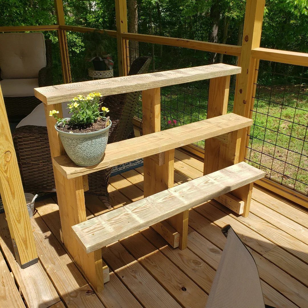 A three-tiered wooden shelf holding a small pot of yellow flowers on a wooden deck.