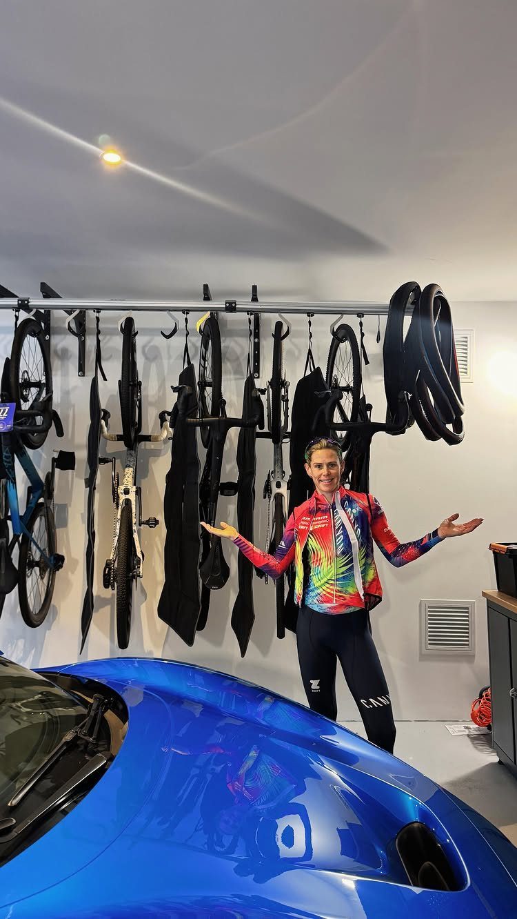 A woman in colorful cycling attire gestures toward a wall-mounted bike track system in a garage.