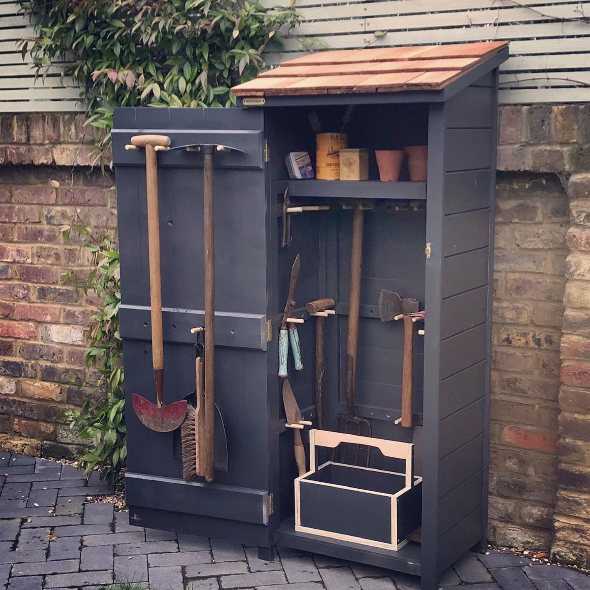 11 Garden Tool Organization Ideas for a Neater Shed or Garage 5 A dark grey vertical wooden garden tool shed standing open against a brick wall.