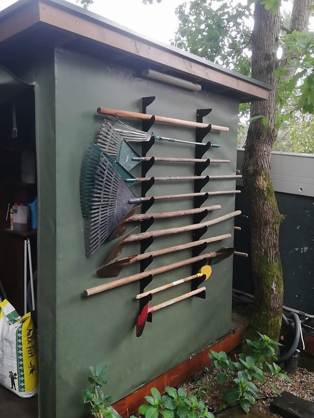11 Garden Tool Organization Ideas for a Neater Shed or Garage 1 Long-handled garden tools hanging on a black wall-mounted rack attached to a green shed.