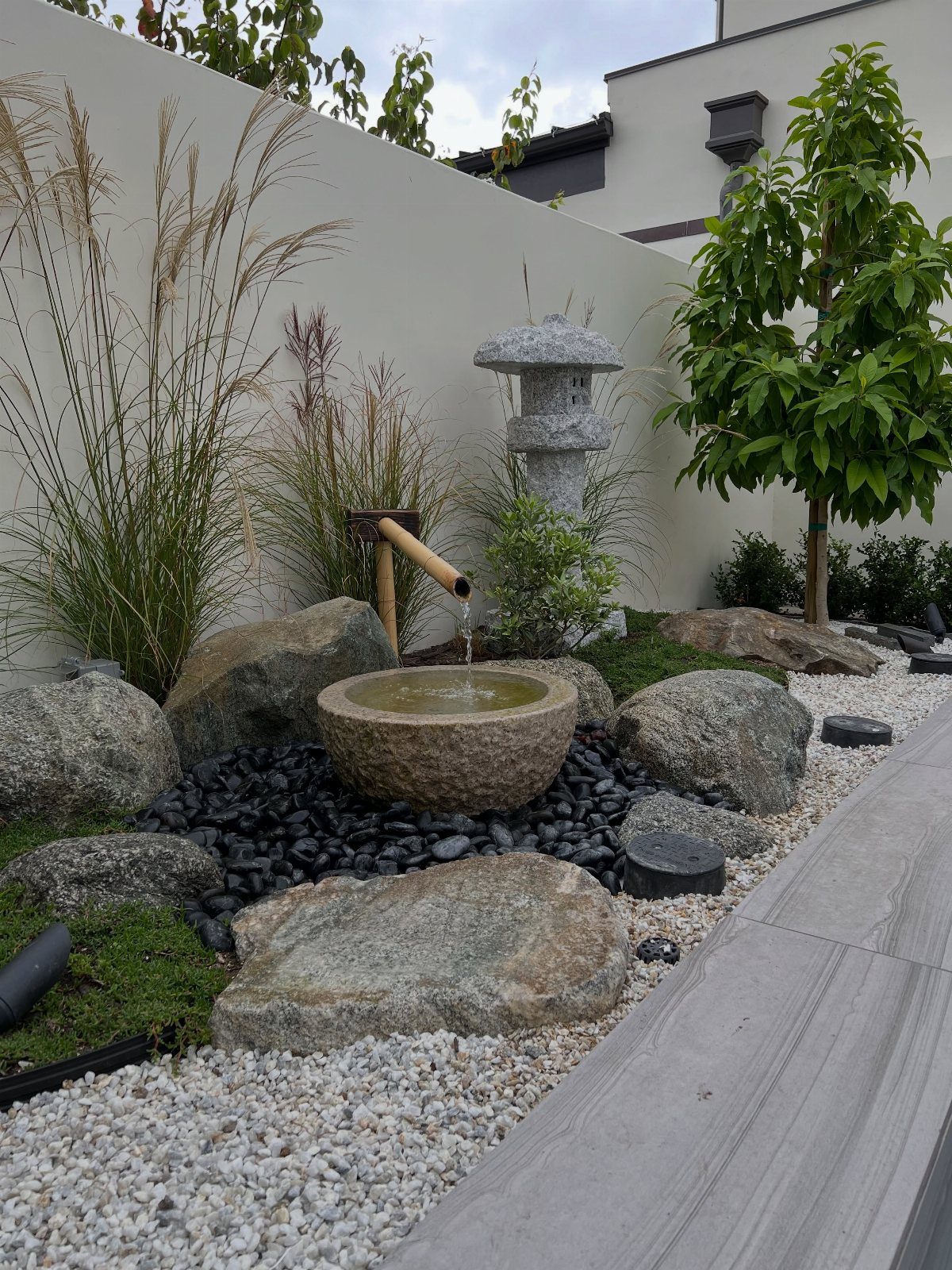 A stone water basin with a bamboo spout surrounded by rocks and white gravel in a Zen garden.